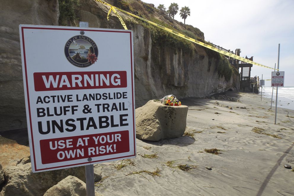 One of several warning signs is posted next to the sand rock debris left from Friday's sea cliff collapse that killed three people near the Grandview Beach access stairway in the beach community of Leucadia, Saturday, Aug. 3, 2019, in Encinitas, Calif. A bouquet of flowers has been placed one of the chunks of sand stone. Officials have reopened much of the Southern California beach on Saturday. (Hayne Palmour IV/The San Diego Union-Tribune via AP)