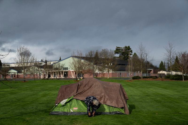 With Fruitdale Elementary School in the background, a homeless man adjusts his shoe at Fruitdale Park, Saturday, March 23, 2024, in Grants Pass, Ore. The rural city of Grants Pass in southern Oregon has become the unlikely face of the nation’s homelessness crisis as its case over anti-camping laws goes to the U.S. Supreme Court. The case has broad implications for cities, including whether they can fine or jail people for camping in public. (AP Photo/Jenny Kane)